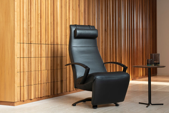 Moderner Relaxsessel von JORI in schwarzem Leder mit hoher Rückenlehne und Nackenrolle – stilvoll platziert vor einer Wand aus warmem Holzlamellen.