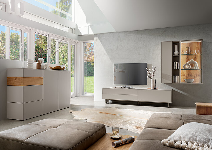 Moderner Wohnraum mit grauem Sofa, Sideboard, TV-Möbel und Wandregal aus dem Wohnprogramm Giorno, große Fensterfront mit Blick ins Grüne.