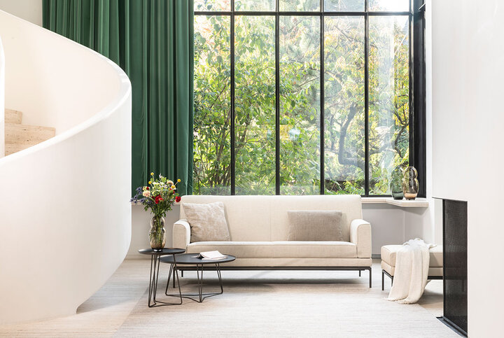 Elegantes Sofa von JORI in hellem Beige – perfekt in Szene gesetzt vor bodentiefem Fenster mit Blick ins Grüne und einem architektonisch geschwungenen Treppenaufgang.