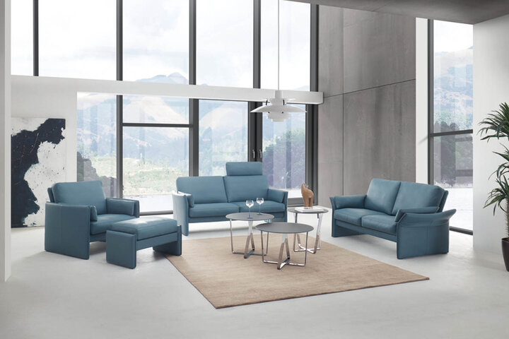 Wohnzimmer mit blauen Ledersofas und Sesseln von Erpo, einem runden Couchtisch und großen Fenstern, die den Blick auf eine bergige Landschaft freigeben.
