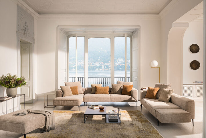Weitläufige JORI-Polstergruppe in Beige mit verschiedenen Modulen, eleganten Metallfüßen und harmonisch abgestimmten Kissen – mit Blick auf den See und hoher Decke im klassischen Interieur.