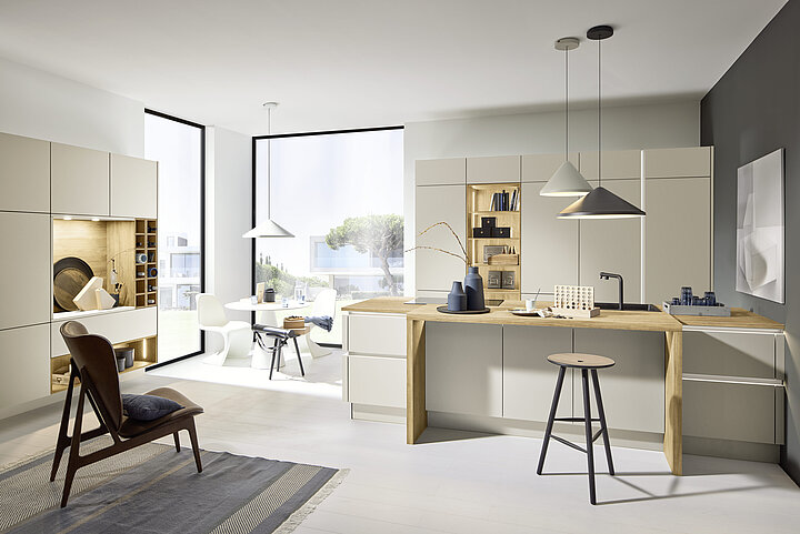 Offene Nolte Küche PERFECT PLUS in Sahara mit beigen Fronten, Holz-Kochinsel und Essbereich vor bodentiefem Fenster.