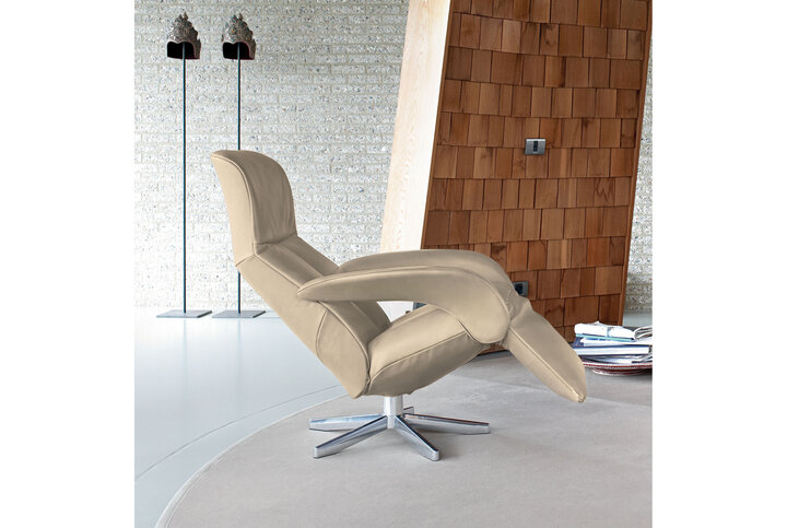 Ergonomischer Relaxsessel von JORI in heller Lederoptik – in der Liegeposition vor moderner Kulisse mit Holzschindelwand und dekorativen Skulpturen.