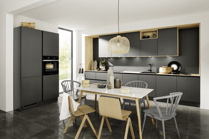 Moderne Nolte Küche mit mattschwarzen Fronten, integriertem Backofen, Holzrahmen an Oberschränken, Esstisch mit gemischten Stühlen und geflochtener Hängeleuchte.