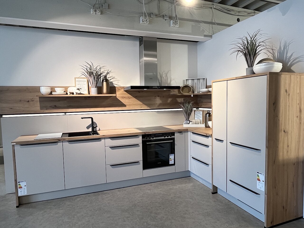 U-Küche in mattem Dunkelgrün mit Holz-Arbeitsplatte in Eichenoptik, schwarzen Griffleisten, Hochglanz-Oberschränken und offenem Regalwürfel-Element, präsentiert in der Ausstellung von Einrichtung Leber.