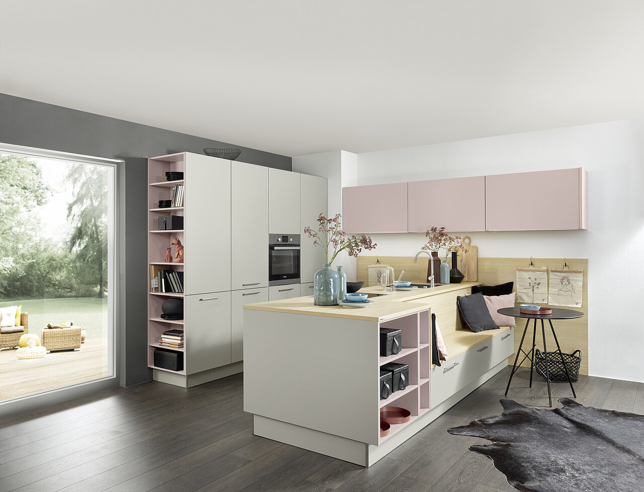Moderne Nolte Küche mit hellen Fronten, rosa Hängeschränken, offener Regalnische, Kücheninsel mit Holzarbeitsplatte und Sitzbank mit Kissen vor weißer Wand und großem Fenster zur Terrasse.