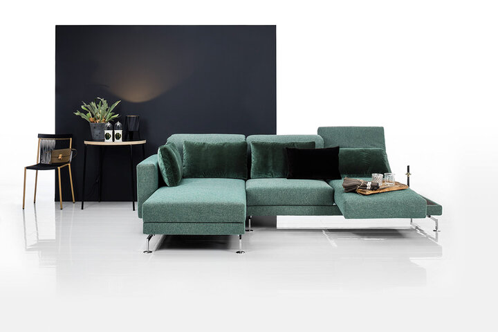 Grünes Stoffsofa von brühl mit verstellbarem Sitzelement und Kissen, vor dunkler Wand.