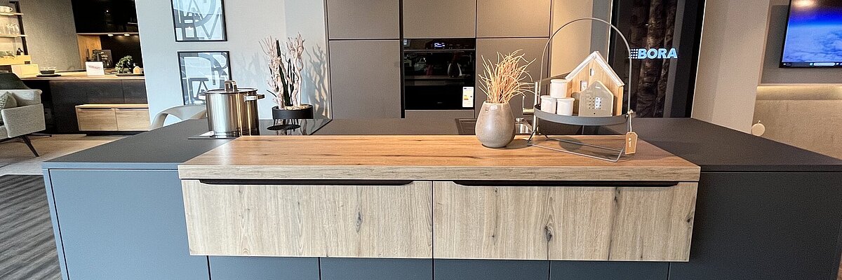 Moderne L-Küche in Cremeweiß mit Holzdekor-Arbeitsplatte, offenen Holzregalen, Edelstahlhaube und hohem Schrankelement, in der Ausstellung von Einrichtung Leber.