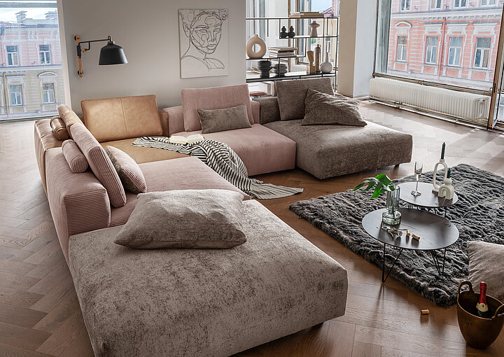 Großzügige, moderne Wohnzimmerecke mit modularer Polstergarnitur in verschiedenen Stoffen, zwei runden Couchtischen auf Teppich und großen Fenstern mit Blick auf Altbauten.