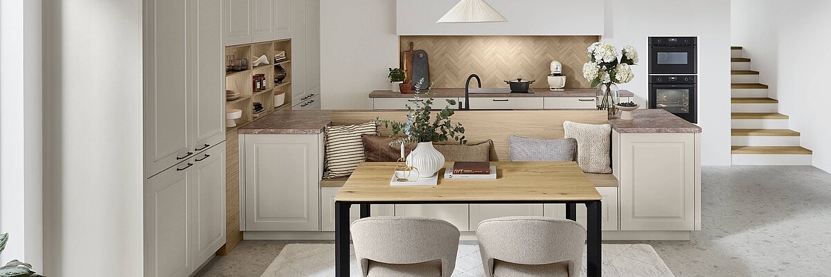 nobilia Landhausküche Sylt 848 in Creme mit kassettierten Rahmenfronten, integrierter Sitzbank und Holz-Esstisch.