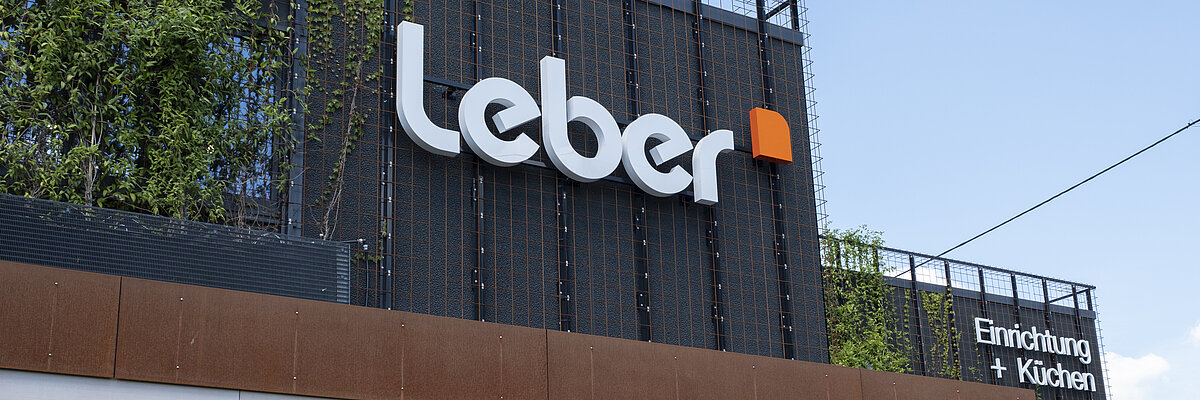 Außenfassade eines Gebäudes mit dem Schriftzug 'leber' und dem Zusatz 'Einrichtung + Küchen' auf schwarzem Hintergrund mit Metallgitter und Pflanzen im oberen Bereich.