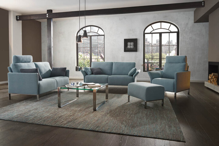 Modernes Wohnzimmer mit blauen Sofas und Sesseln von Erpo, Glas-Couchtisch und Blick durch große Fenster auf eine städtische Landschaft.