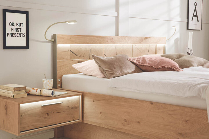 Schlafzimmer mit Holzbettrahmen von Casada, beleuchtetem Kopfteil, Dekokissen und einem Nachttisch, auf dem Bücher und eine Tasse stehen. Ein Poster an der Wand ist sichtbar.
