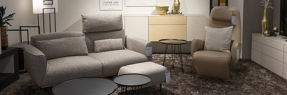 Headerbild: Wohnzimmer mit grauem Sofa, beigem Sessel, zwei runden Couchtischen und Stehlampe auf braunem Teppich.